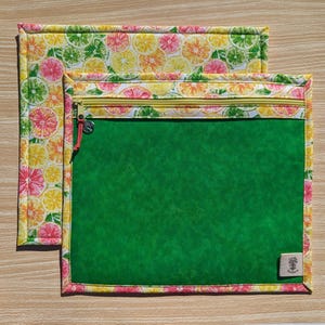 May include: Two rectangular fabric pouches. The top pouch is open, and the bottom pouch has a zipper closure. Both have a bright green center and a border with a colorful citrus fruit pattern. The zipper is gold-colored, and the zipper pull has a small charm.
