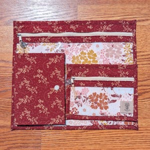 May include: A square fabric organizer featuring a deep red background with a white floral pattern. It includes a top zippered compartment, a left-side flap pocket with a snap closure, and a right-side zippered pocket with a small rectangular label.
