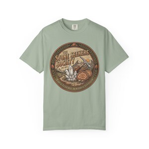 Könnte beinhalten: Ein salbeigrünes T-Shirt mit einem kreisförmigen Aufdruck mit dem Text THE STONE SEEKERS SOCIETY EST. 1974 und Every Trail Has A Hidden Treasure. Das Motiv zeigt einen Geologenhammer, Kristallcluster und Gesteinsformationen.