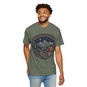 May include: An olive green t-shirt featuring a graphic print of a motorcycle parked near a tent in a mountain forest at night. The design includes a crescent moon, stars, and the text "Where The Road Ends Adventure Begins" in a shield shape.