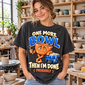 以下が含まれることがあります： チャコールグレーのTシャツ。顔のついたオレンジ色のボウルが青い陶芸ろくろにもたれかかっているグラフィックが描かれています。テキストは黄色と青の文字で「ONE MORE BOWL THEN I'M DONE PROBABLY」と書かれています。半袖のクルーネックTシャツです。