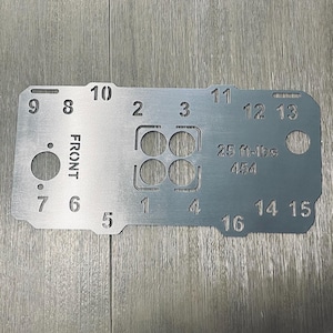 May include: A silver metal tool with numbers 1-16, the word "FRONT", and the measurement "25 ft-lbs 454" etched on its surface. The tool has several holes and cutouts.