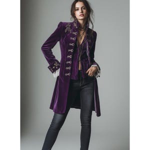 May include: A long, purple velvet coat with gold embroidery on the shoulders and cuffs. The coat has decorative buttons down the front and lace trim at the sleeves. The model is wearing black jeans.