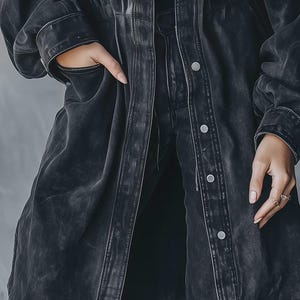 May include: A long, dark grey denim jacket with a button-up front and pockets. The jacket has a worn, distressed look, and the sleeves are long. The model is wearing the jacket with matching denim trousers.