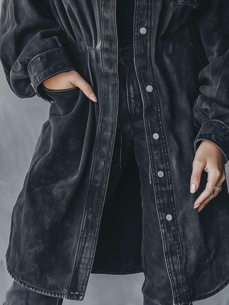 May include: A long, dark grey denim jacket with a button-up front and pockets. The jacket has a worn, distressed look, and the sleeves are long. The model is wearing the jacket with matching denim trousers.