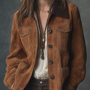 Puede incluir: Chaqueta de ante marrón con cuello marrón oscuro y cierre de botones. La chaqueta tiene dos bolsillos en el pecho y dos bolsillos laterales. La modelo lleva una blusa blanca y pantalones marrones. Se ve un collar largo de cuentas.