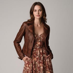 Op de afbeelding: Bruine leren jas met een klassiek asymmetrisch ritssluiting. De jas wordt gedragen over een jurk met bloemenprint in tinten rood, bruin en crème. Het model staat voor een neutrale achtergrond.