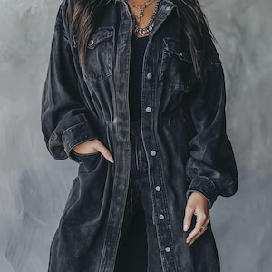 May include: A long, dark gray denim jacket with a button-down front, two chest pockets, and a cinched waist. The jacket has long sleeves and is worn over a black top. The model is wearing layered necklaces.