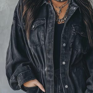 May include: A black denim jacket with two chest pockets and silver buttons. The jacket has a classic collar and a slightly faded appearance. The model is wearing a black top and layered necklaces.