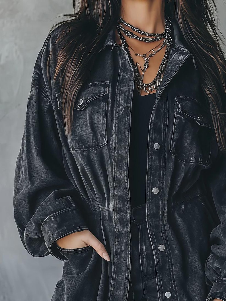 May include: A black denim jacket with two chest pockets and silver buttons. The jacket has a classic collar and a slightly faded appearance. The model is wearing a black top and layered necklaces.