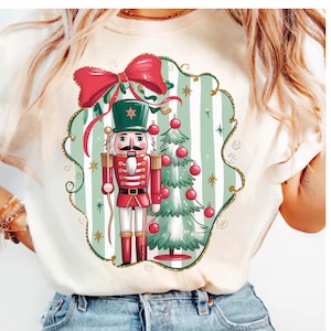 May include: Cream-colored t-shirt featuring a Christmas-themed graphic. The design includes a red and green nutcracker soldier, a Christmas tree with red ornaments, and a large red bow. The graphic is set against a striped background.
