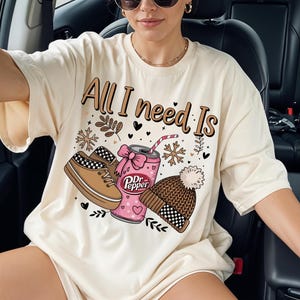 Könnte beinhalten: Cremefarbenes T-Shirt mit dem Text "All I need Is" und Illustrationen eines Schuhs, einer Dr. Pepper-Dose und einer Mütze. Der Schuh hat ein schwarz-weißes Schachbrettmuster. Die Dr. Pepper-Dose hat eine rosa Schleife.