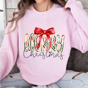 May include: A light pink sweatshirt with the words "MERRY Christmas" in a festive design. The word "MERRY" is in a colorful plaid pattern, topped with a red bow. The word "Christmas" is in a cursive font.