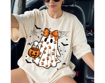 Könnte beinhalten: Cremefarbenes T-Shirt zum Thema Halloween mit Geisterdesign. Der Geist hat eine orange Schleife, trägt einen Kürbis-Eimer und ist mit Kürbis- und Spinnennetz-Grafiken verziert. Fledermäuse und Sterne vervollständigen das festliche Design.
