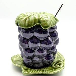 May include: A ceramic jar shaped like a blackberry, with a green leaf-shaped lid and base. The jar is a deep purple color, with a glossy finish. A small metal spoon rests in the jar. The base is a green leaf shape.