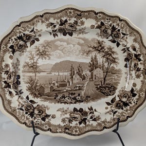 May include: An oval ceramic platter featuring a brown and white transferware landscape scene of a harbor with buildings, trees, and hills. The scalloped rim is decorated with a detailed border of floral patterns and foliage in matching brown tones.