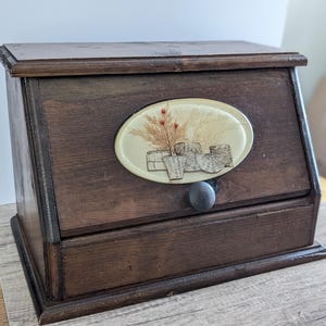May include: A dark brown wooden bread box with a slanted front door featuring a round black knob. An oval-shaped cream-colored inlay is centered on the door, displaying a detailed illustration of wheat stalks and woven baskets in brown tones.