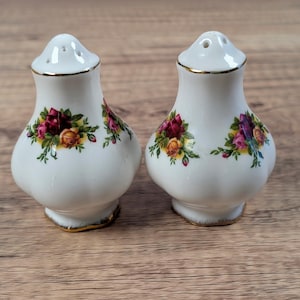 May include: A pair of white ceramic salt and pepper shakers with a bulbous shape and gold-trimmed rims. Each shaker features a colorful floral pattern consisting of red, yellow, and pink roses with green leaves printed on the front and sides.