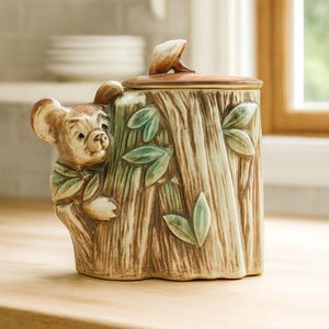 May include: A ceramic cookie jar shaped like a tree stump with a bear cub peeking out. The jar is painted in shades of brown and green, with a lid featuring a small bird. The design includes leaves and tree bark details, creating a whimsical, rustic aesthetic.