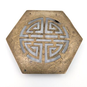 May include: Hexagonal brass trivet with a central cutout design featuring a stylized symbol. The trivet has floral and leaf patterns around the edges. The brass has a warm, aged patina.