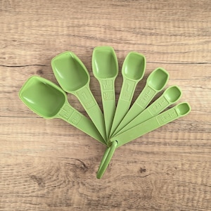 May include: A set of six light green measuring spoons fanned out on a wooden surface. Each spoon has a different size, with measurements printed on the handles. The spoons are made of plastic and are a solid, uniform color.