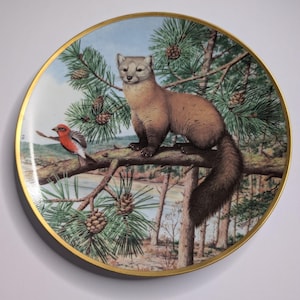 May include: Decorative plate featuring a detailed illustration of a pine marten perched on a tree branch with a small bird. The plate has a gold rim and a nature-themed design with a forest and lake background.