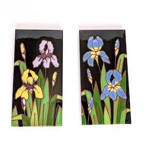 May include: Two rectangular decorative tiles featuring iris flowers. One tile showcases yellow and purple irises, while the other displays blue and yellow irises. The flowers are set against a black background with green leaves.