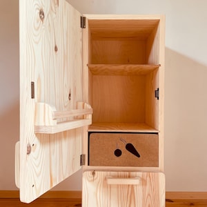 Könnte beinhalten: Ein helles Holzschrank mit einer offenen Tür, die Regale und eine Schublade freigibt. Der Schrank hat eine natürliche Holzmaserung. Eine Kiste mit einer Karotten- und Zwiebelgrafik ist sichtbar. Der Schrank ist für die Aufbewahrung konzipiert.