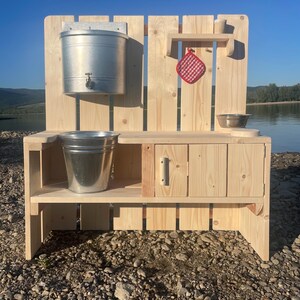Pode incluir: Uma cozinha de brincar de madeira para exterior com acabamento natural. Possui um balde de metal com torneira, uma prateleira com um pega panelas xadrez vermelho e branco e uma tigela pequena. Um segundo balde de metal fica embaixo.