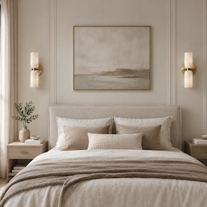 May include: A bedroom scene featuring a neutral-toned landscape painting in a thin frame centered above a bed with a beige upholstered headboard. Two brass wall sconces with cylindrical frosted glass shades flank the artwork on both sides.