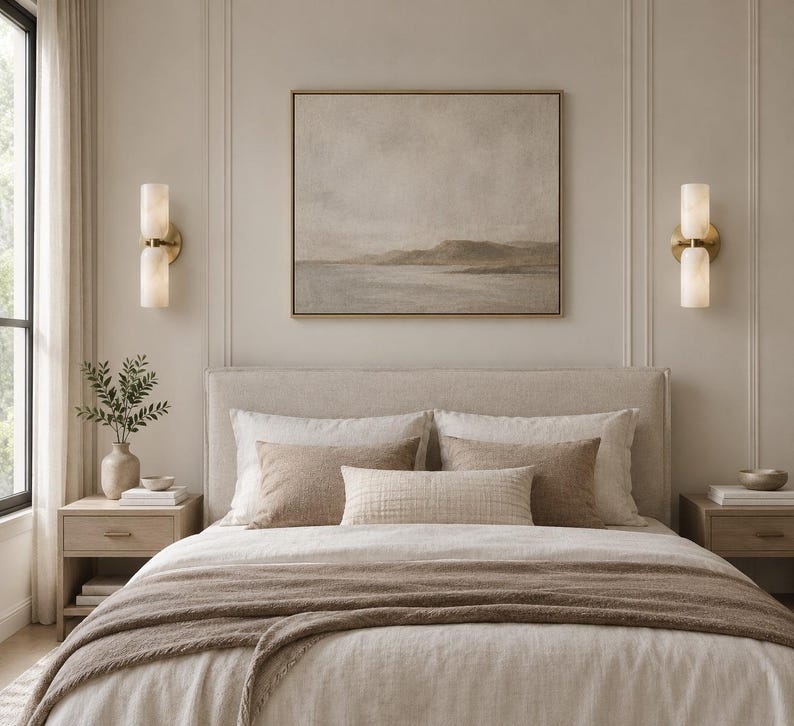 May include: A bedroom scene featuring a neutral-toned landscape painting in a thin frame centered above a bed with a beige upholstered headboard. Two brass wall sconces with cylindrical frosted glass shades flank the artwork on both sides.