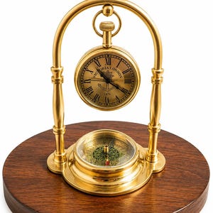 May include: A brass desk clock and compass set mounted on a circular dark wood base. The clock features a gold face with Roman numerals, the text "VICTORIA LONDON" and "1876," and an anchor graphic. A brass arch supports the hanging clock.