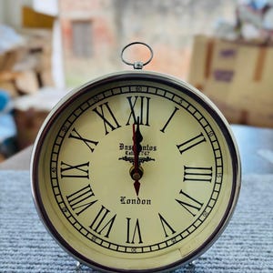 Puede incluir: Un reloj redondo plateado con esfera crema y números romanos negros. La esfera del reloj muestra el texto "Basontos Est. 747 London". Tiene un pequeño asa en la parte superior y está sobre una superficie gris.