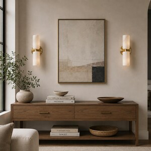 May include: A rectangular wooden console table with two drawers and a lower shelf, featuring a framed abstract painting, two cylindrical wall sconces, a ceramic vase with greenery, two bowls, and four stacked books titled Live Beautifully, This Is Home, The Kinfolk Home, and Vogue.