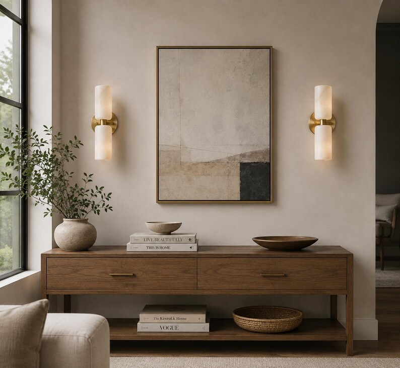 May include: A rectangular wooden console table with two drawers and a lower shelf, featuring a framed abstract painting, two cylindrical wall sconces, a ceramic vase with greenery, two bowls, and four stacked books titled Live Beautifully, This Is Home, The Kinfolk Home, and Vogue.
