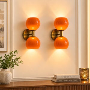 May include: Two wall-mounted light fixtures featuring a brass-toned circular base and a central rod holding two spherical orange glass shades. Each lamp emits a warm glow, with one shade pointing upward and the other downward, creating a symmetrical effect.