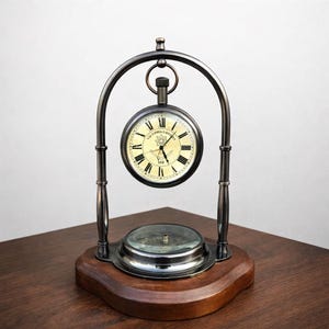 May include: Antique-style clock with a cream-colored face, black Roman numerals, and the text "Victoria London". The clock is suspended within a dark metal frame, resting on a wooden base with a compass.