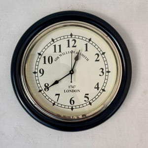 May include: A circular wall clock with a dark blue frame and a cream face. The clock displays black Roman numerals, black hands, and the text "SIR WILLIAM & SMITH LONDON 1747". It has a silver-coloured inner rim.