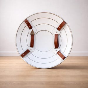 May include: A white, round shield with concentric silver lines and brown leather straps. The shield has a silver rim and is designed with a classic, superhero aesthetic. The straps are riveted to the shield.