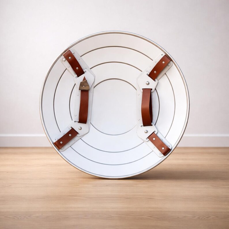 May include: A white, round shield with concentric silver lines and brown leather straps. The shield has a silver rim and is designed with a classic, superhero aesthetic. The straps are riveted to the shield.