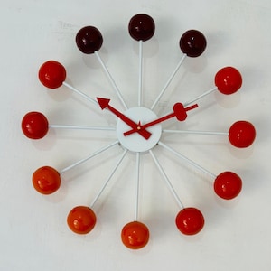 Puede incluir: Un reloj de pared moderno con una esfera blanca y manecillas rojas. El reloj presenta radios blancos con esferas rojas y granates que marcan las horas. El diseño del reloj es una adición única y elegante a cualquier habitación.