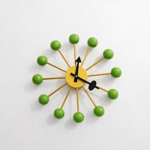 May include: A modern wall clock with a yellow center and gold-colored spokes. Each spoke ends with a green sphere. The clock hands are black. This decorative clock is a unique piece of home decor.