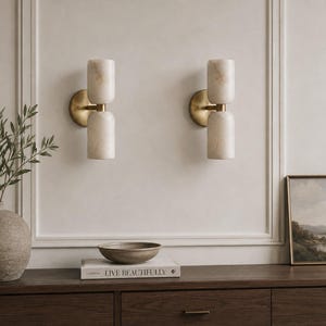 May include: Two wall sconces featuring cylindrical, cream-colored stone shades with visible veining, mounted on circular brass-toned backplates. Each fixture consists of two stacked stone cylinders connected by a central brass-toned horizontal arm.