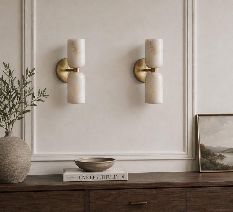 May include: Two wall sconces featuring cylindrical, cream-colored stone shades with visible veining, mounted on circular brass-toned backplates. Each fixture consists of two stacked stone cylinders connected by a central brass-toned horizontal arm.