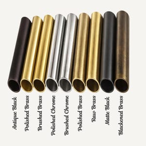 May include: Nine cylindrical metal tubes arranged in a row, showcasing various finishes labeled as Antique Black, Polished Brass, Brushed Brass, Polished Chrome, Brushed Chrome, Polished Brass, Raw Brass, Matte Black, and Blackened Brass.