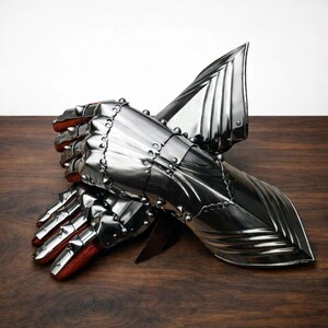 May include: Pair of polished steel gauntlets with riveted construction and red leather lining. The gauntlets feature articulated fingers and flared cuffs, designed for medieval armor. The metal has a reflective surface, and the gauntlets are displayed on a wooden surface.