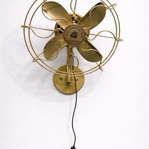 May include: A vintage-style brass wall fan with four curved blades and a protective cage. The fan is mounted on a round base and has a black power cord. The center of the fan reads "MADE FOR ROYAL NAVY LONDON".