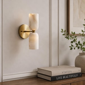 May include: A wall-mounted light fixture featuring a circular brass backplate and a horizontal arm supporting two vertically stacked, cylindrical white stone shades with subtle beige veining. The fixture emits a soft, warm glow from both shades.