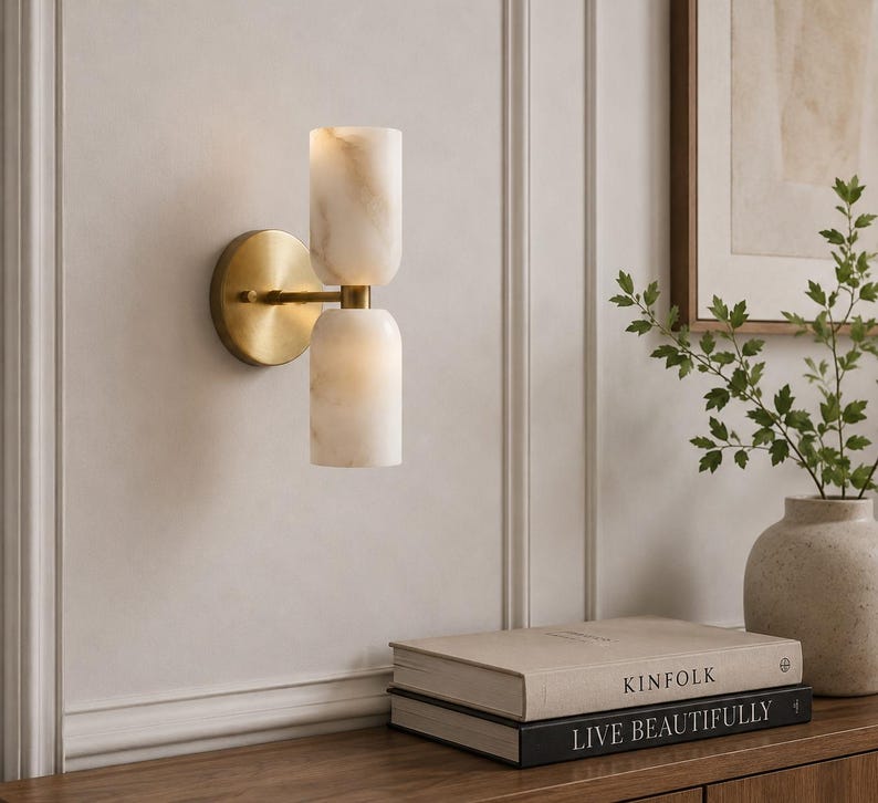 May include: A wall-mounted light fixture featuring a circular brass backplate and a horizontal arm supporting two vertically stacked, cylindrical white stone shades with subtle beige veining. The fixture emits a soft, warm glow from both shades.