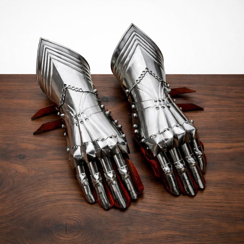 May include: A pair of polished steel gauntlets, featuring articulated fingers and a layered design. The gauntlets have a red interior and are displayed on a wooden surface. The medieval-style armor is designed for protection and display.